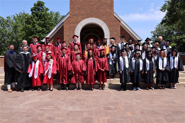 A large group of graduates and speakers post together and smile at the camera from the steps in front of Hawkins Chapel.
